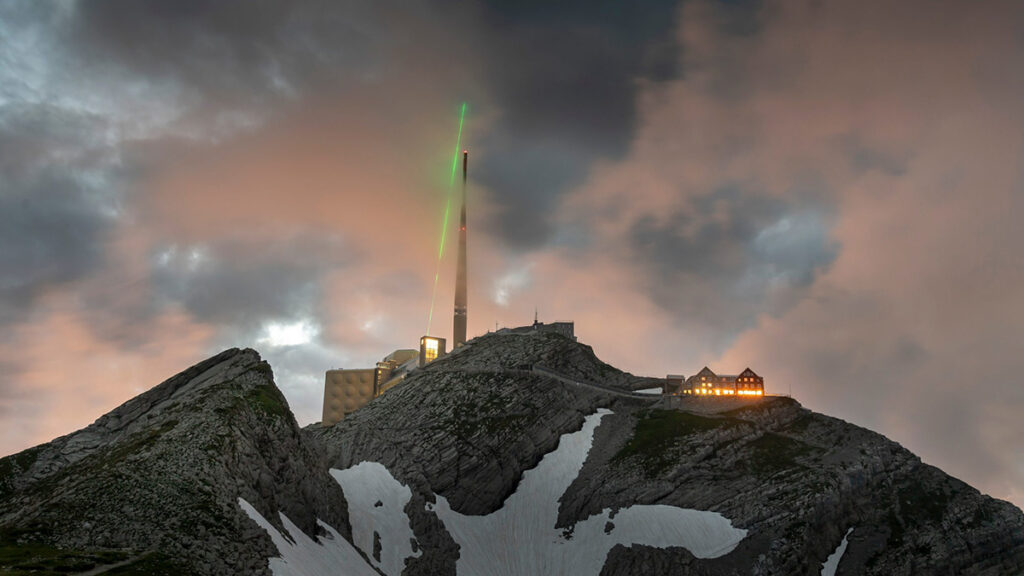The laser device sits on top of Mount Säntis, shooting a green laser into a cloudy sky.