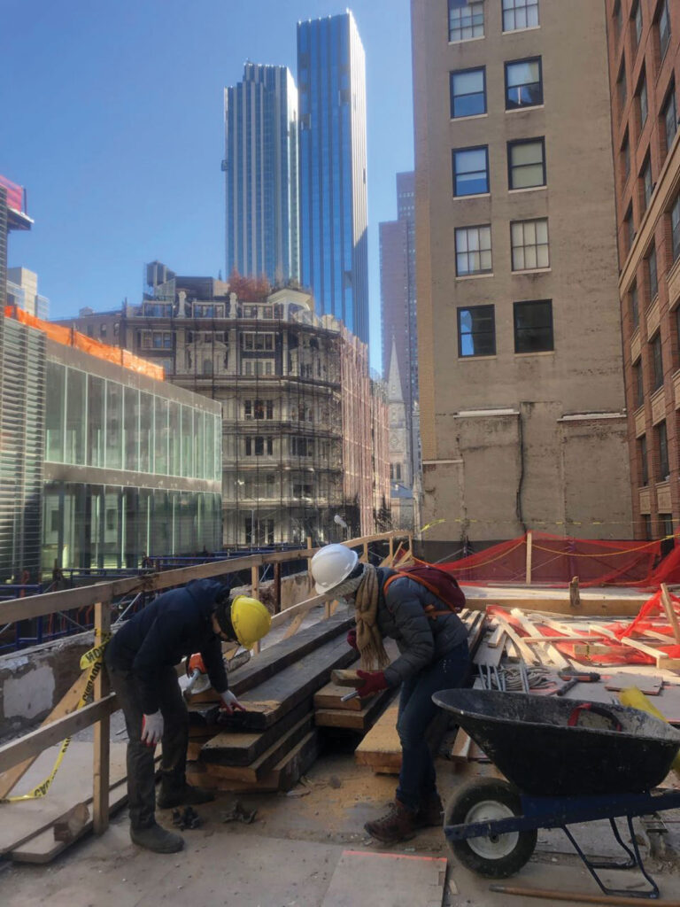 Two people in hard hats inspect timber on the roof of a building with skyscrapers in the background.