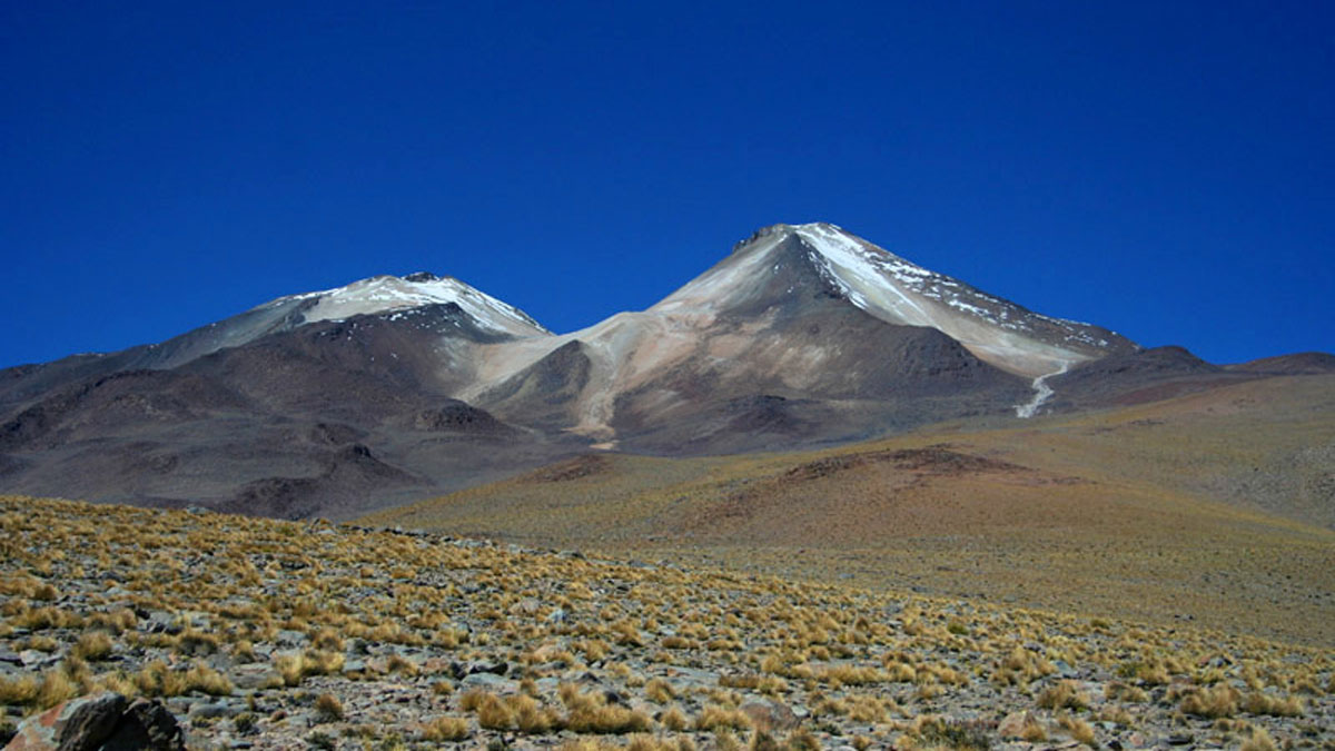 A volcano with two snow-dusted peaks in an arid landscape with a cloudless blue sky.