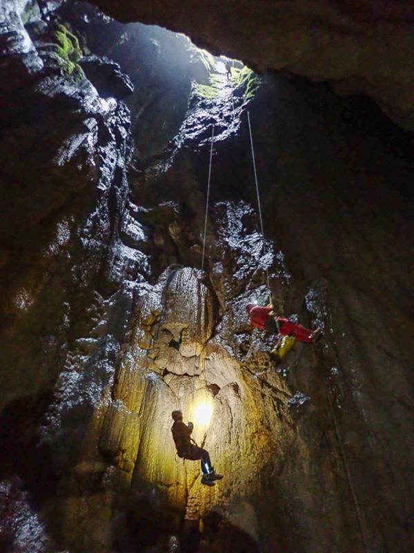 Two people are hanging on ropes down a dark, narrow cave shaft. One wall is illuminated by a lamp.