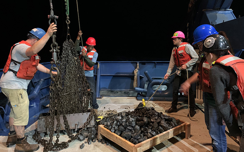 Several people wearing helmets and orange life preservers unload rocks from a chain metal dredge basket at night on the deck of a ship.