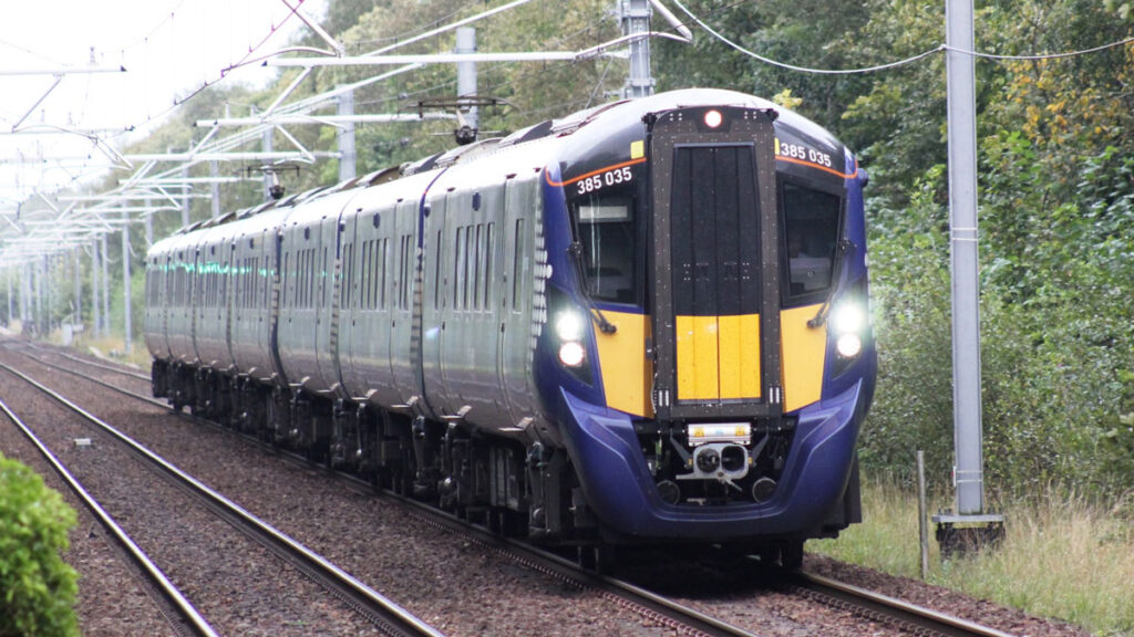 A blue and yellow electrical passenger train travels past green trees toward the viewer along one of two sets of parallel tracks.