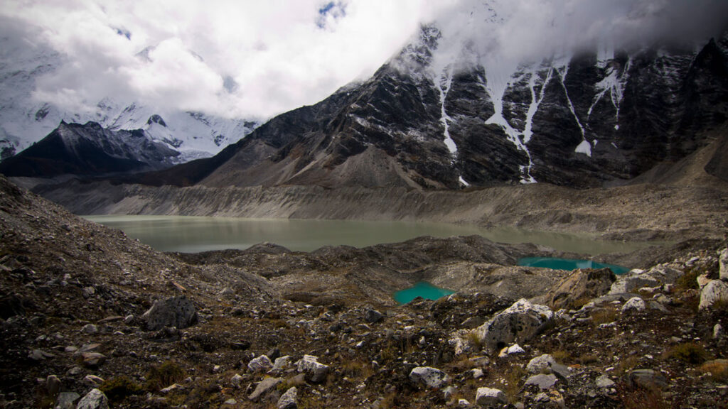 A greenish lake sits in front of a snow-topped mountain under a cloud-filled sky.