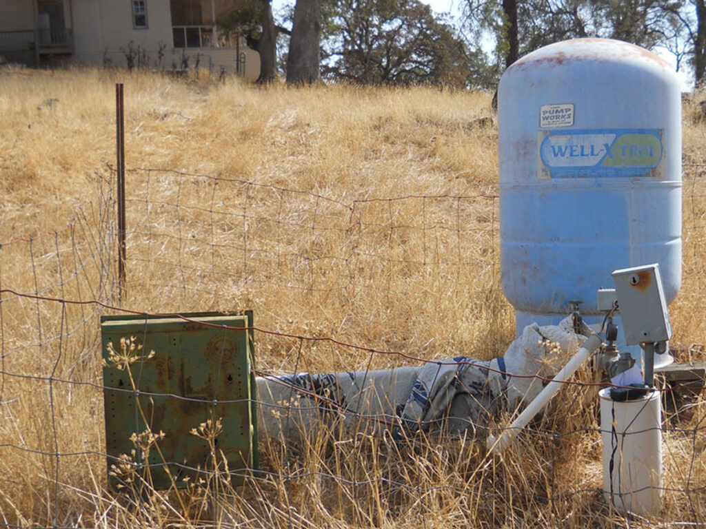 A metal cylinder and box surrounded by dry grass in front of a house in a rural area.