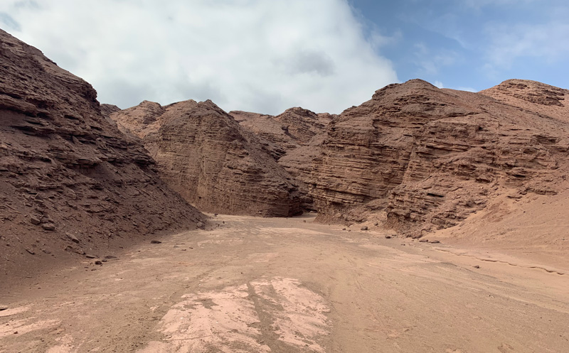 Vista de un cuerpo de roca estratificado de color rojo con una planicie arenosa al centro.