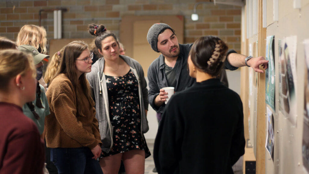 A student points at and discusses a poster hanging on a wall while other students listen and look at the poster.