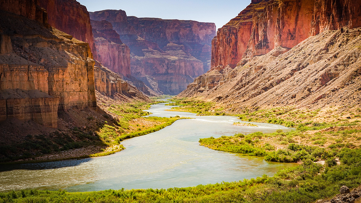 A Sun-speckled river winds through banks populated by green scrub brush. Along the edge of the vegetation, red-brown sand abuts mesas that stretch toward a bright blue sky.