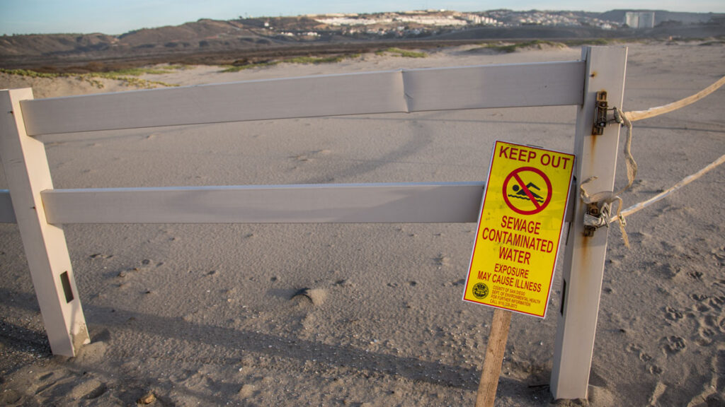 Bright yellow beach closure sign that says “Keep out. Sewage contaminated water. Exposure may cause illness.” The sign is leaning against a gate outside of Border Field State Park, which is located in Imperial Beach, Calif.