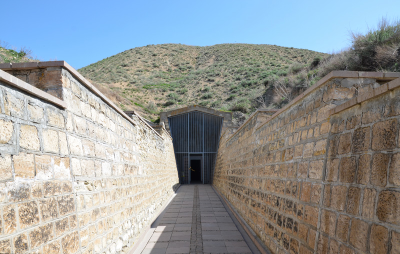 A tall door surrounded by bars is at the end of a long, narrow brick walkway. The doorway leads into a hill covered in dirt and low green vegetation.