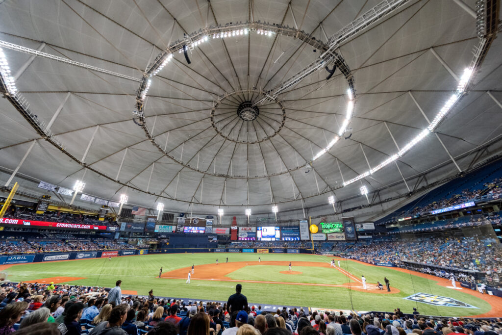 This photograph shows the interior of a domed baseball stadium. The gray dome is closed during this game. The field is photographed from behind the third-base dugout and looks out past the pitcher’s mound toward right field.