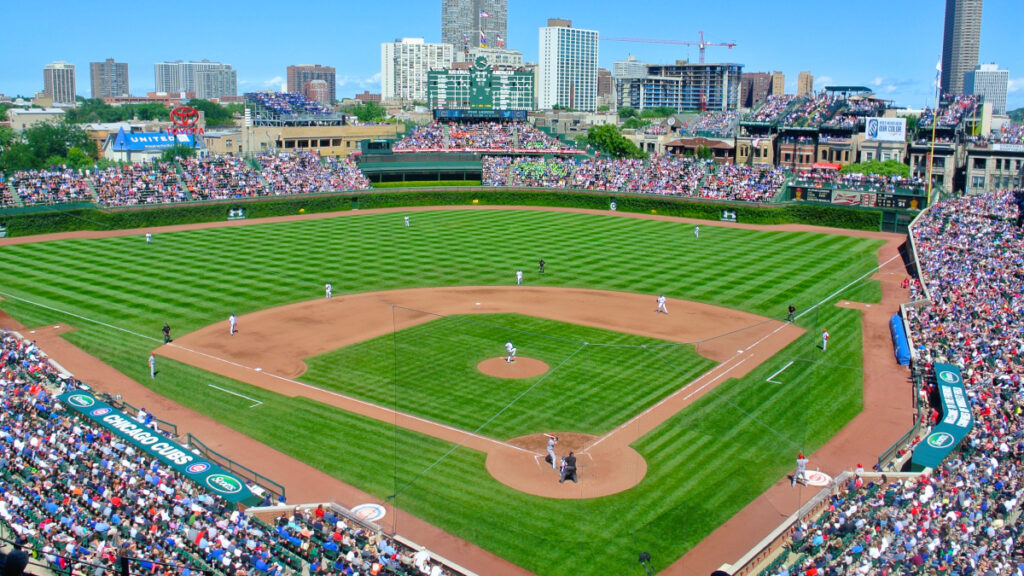 A packed baseball stadium with a bright green field on a sunny day. The field is photographed from above and behind home plate looking toward the outfield.
