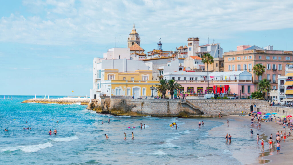 Beachgoers swim and lounge seaside on a calm day on a Catalan beach. The waters are a clear light blue, the sky is partly cloudy, and colorful buildings fill the background.
