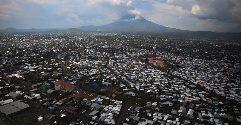 Photo of many white-roofed buildings in the fore- and mid-ground and a mountain surrounded by clouds in the background.