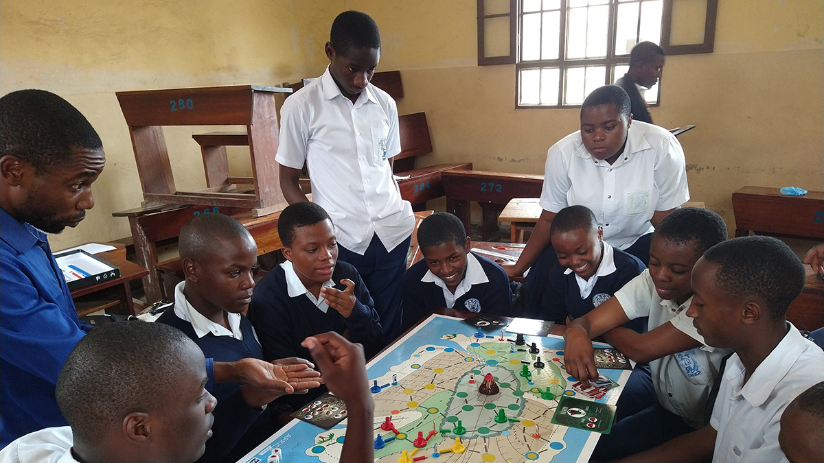 A group of teenagers in white and blue uniforms sits around a board game with colorful pieces.
