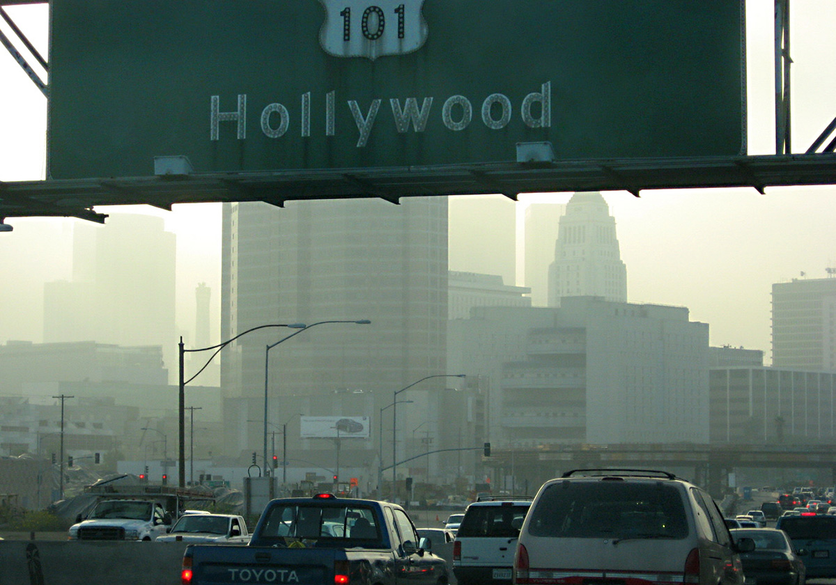 Hollywood road sign hangs above cars along Highway 101. In the distance is the smog-filled skyline of Los Angeles.