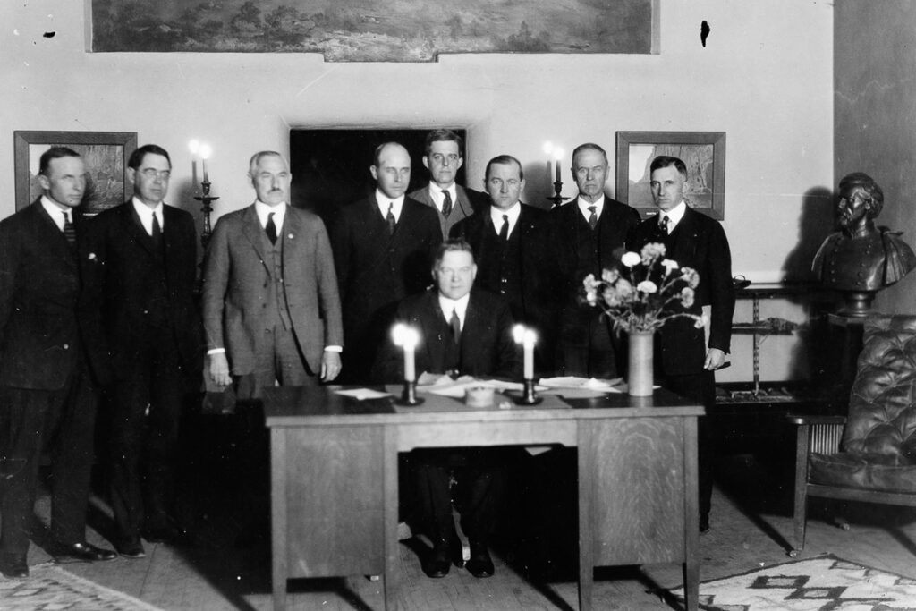 Grainy black and white photo of eight men in suits standing behind then-U.S. Secretary of Commerce Herbert Hoover, who is seated behind a large wooden desk.