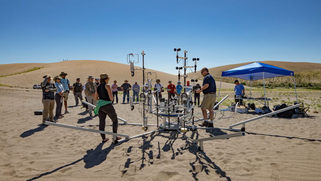 Roughly 20 people stand amid sand dunes under a clear blue sky and near a metal framework equipped with scientific instruments.