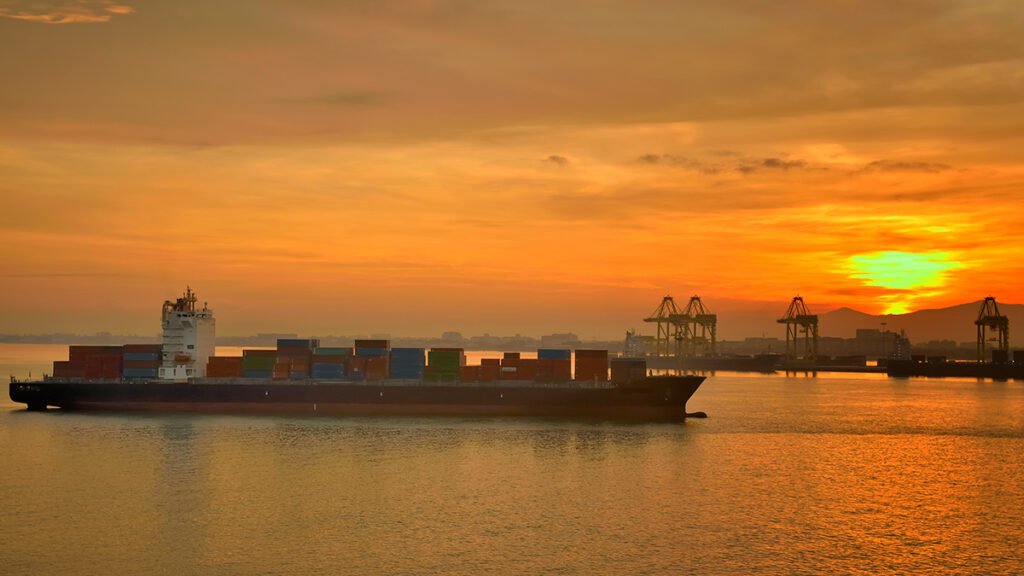 A large boat loaded with shipping containers sails in front of a port as the Sun rises in the background.