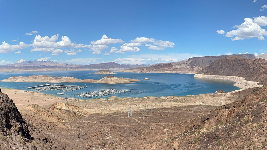 A view of two marinas at the edge of a lake in the distance, with the arid rocky landscape sloping down to the lake in the foreground