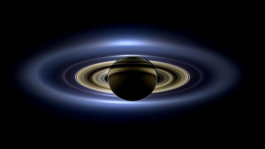 An image of a dark brown sphere and its rings is backlit against a black background. The closer rings glow in yellow-orange and are haloed by the dusty ring system in blue-white. Several bright specks are visible.