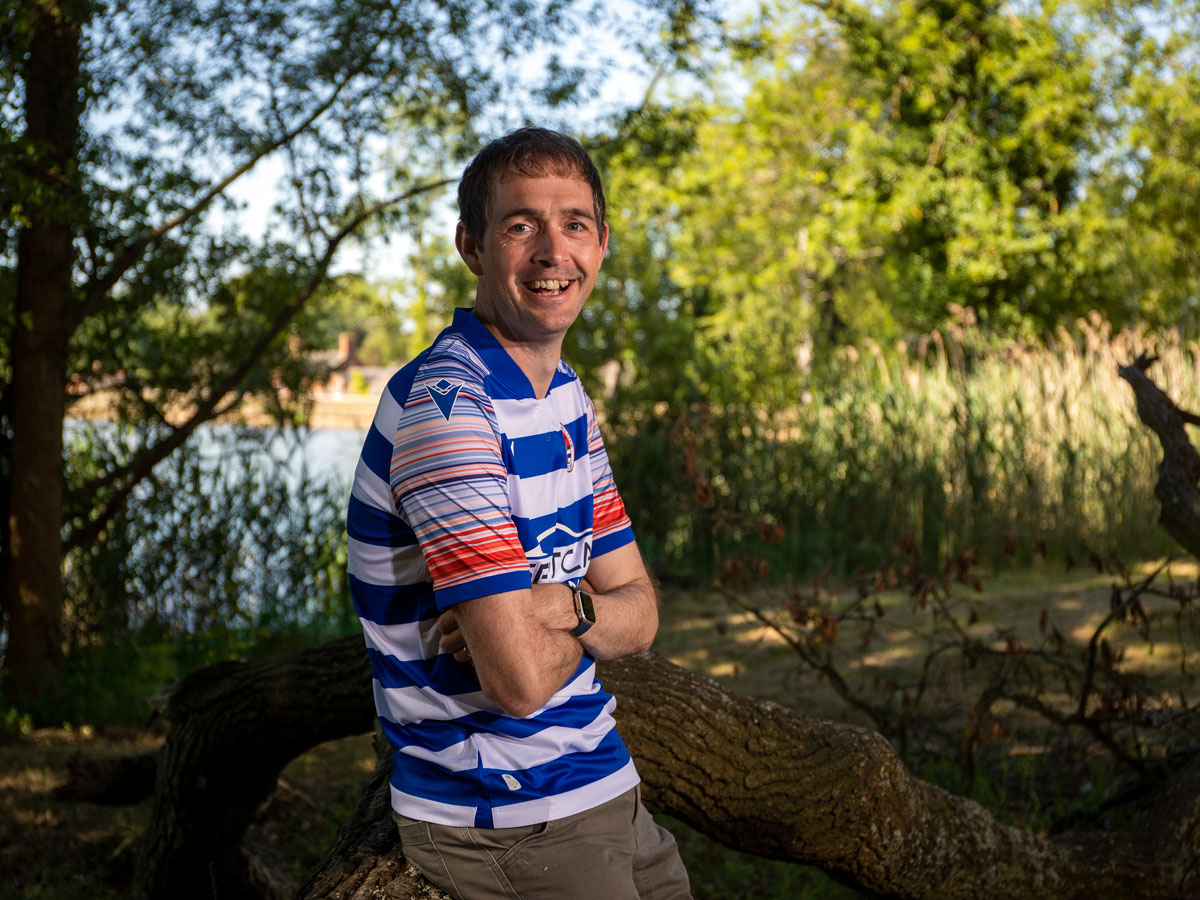 A person wearing a fitted blue and white striped shirt with blue and red stripes on the sleeves is leaning on a log while looking at the camera.