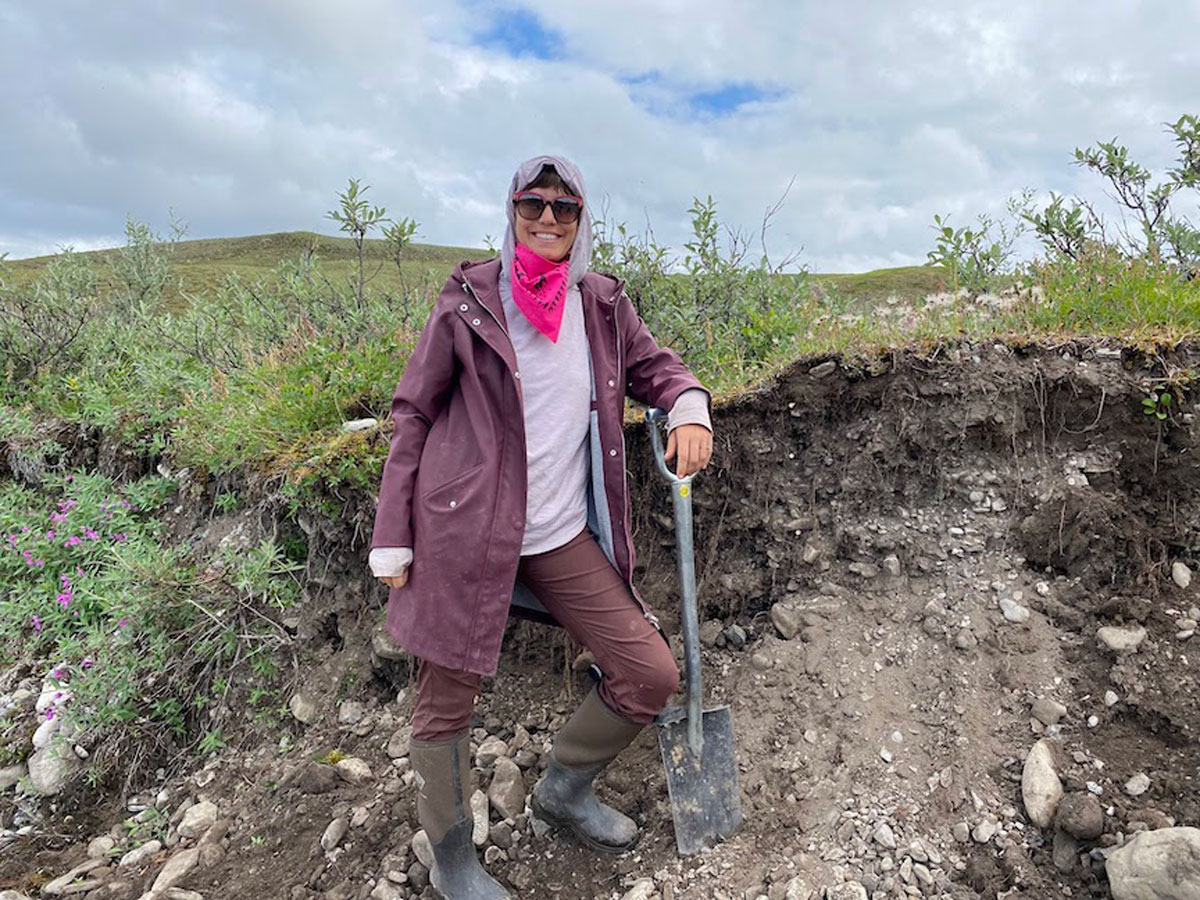 A person wearing sunglasses is looking at the camera and leaning on a shovel next to an outcrop of soil. A green landscape is in the background.