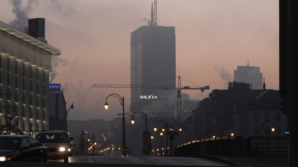 A photo of city buildings at dusk with cars driving by. The air is hazy.