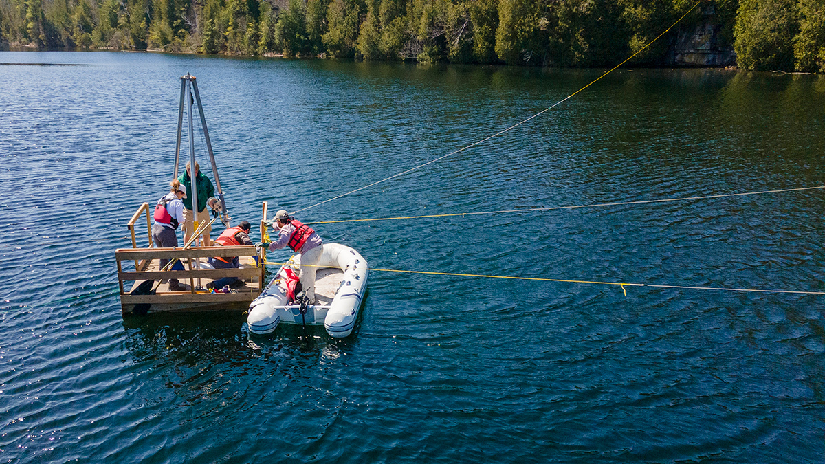 Three people stand on a wooden platform in a lake, and a white, inflatable boat next to the platform holds another person. A 3-meter-tall metal tripod extends above the platform.