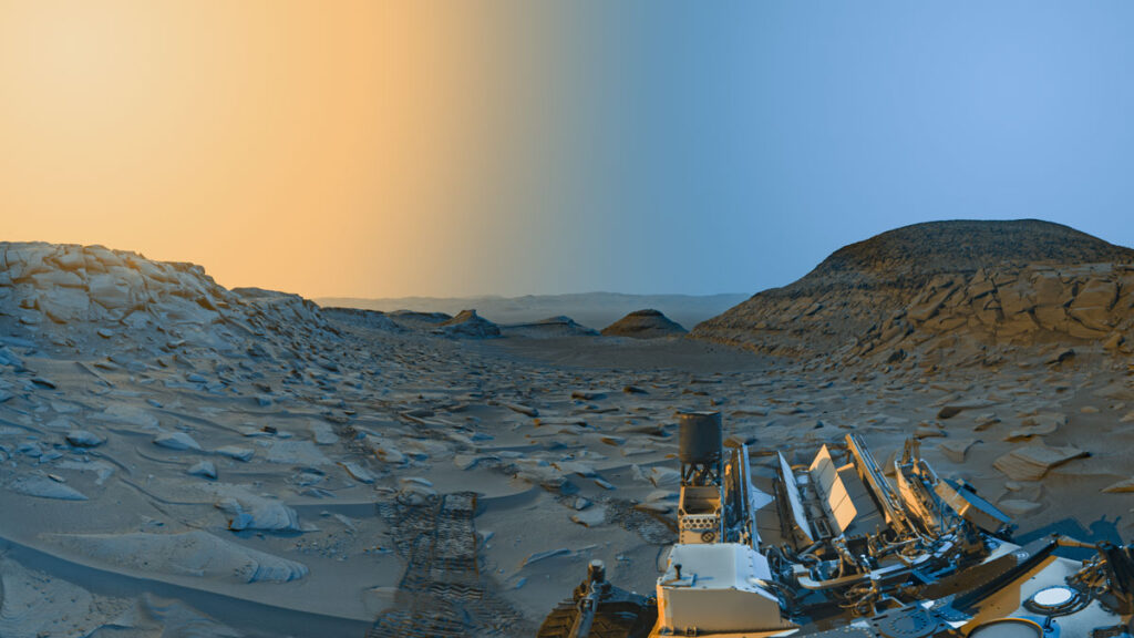 An artificially colorized view of a rocky valley between two hills on Mars, with part of the Curiosity rover visible at bottom. The right portion of the image, under blue sky, represents the scene in the morning, and the left portion, under yellow sky, the afternoon.