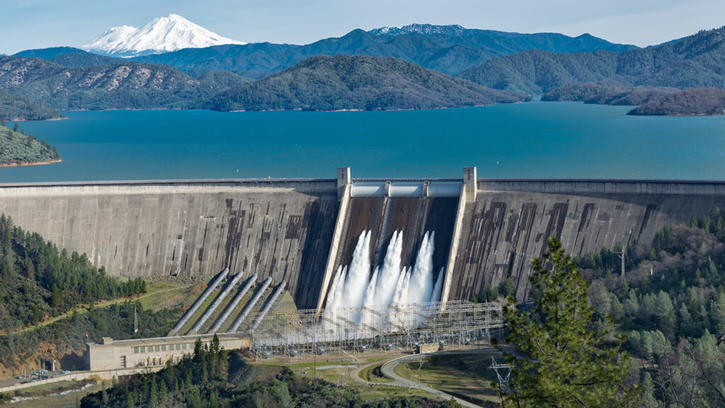 A hydroelectric dam has a very full reservoir at the top and seven release valves open. The reservoir is full of blue-green water, and there are snowcapped mountains in the background.