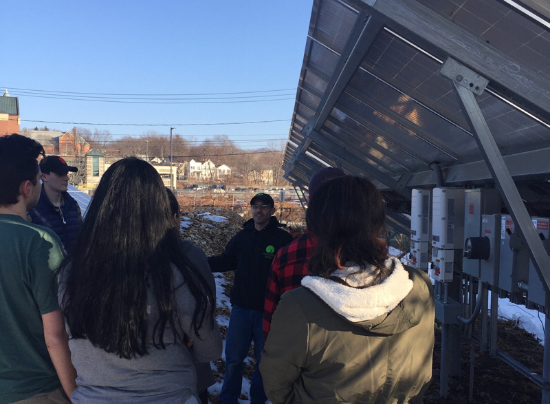 Students standing outside in the shadow of a solar panel installation listen to a tour guide.
