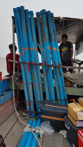 Photo in a small boat at sea. Two people are in the background. In the foreground, a series of 2-meter-long blue tubes stand upright.