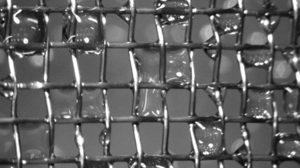 A silver wire mesh collects droplets of water in front of a gray background.