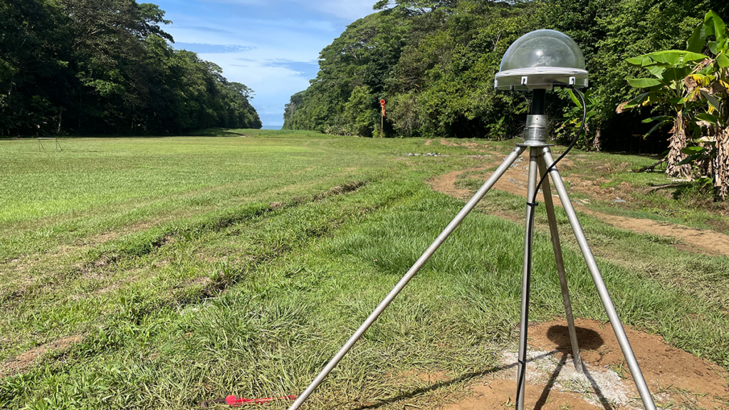 A scientific monitoring instrument with a dome-shaped cover mounted on a tripod sits in a grassy strip of land between two forested areas.