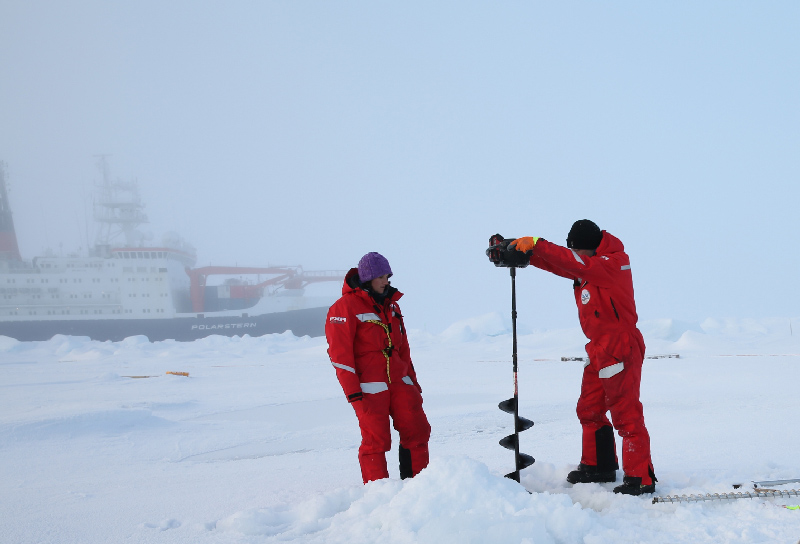 Two expedition members use an auger to drill a hole into sea ice to install an autonomous observatory.