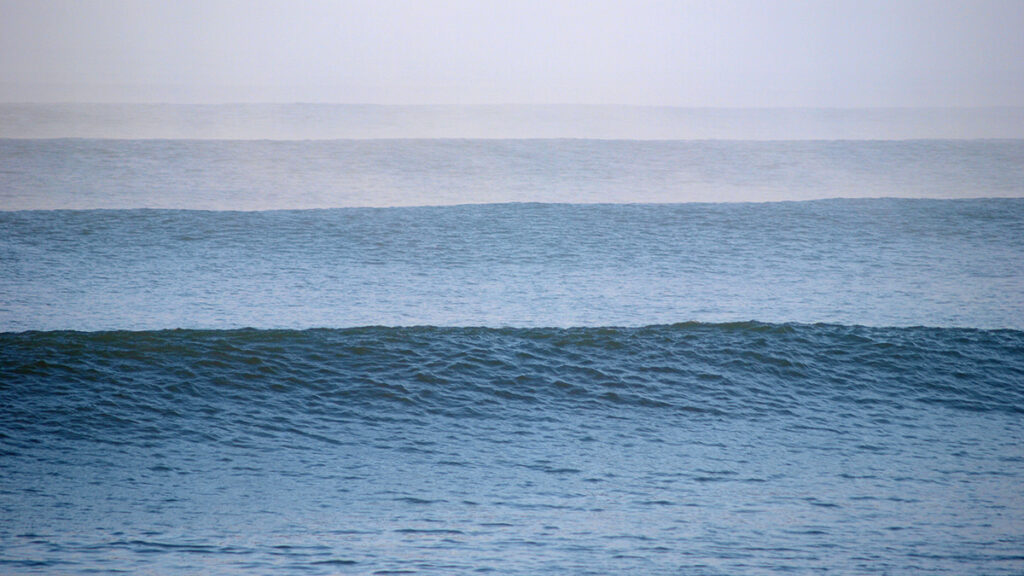 An ocean wave prepares to break, while other waves close in behind it under a hazy sky.