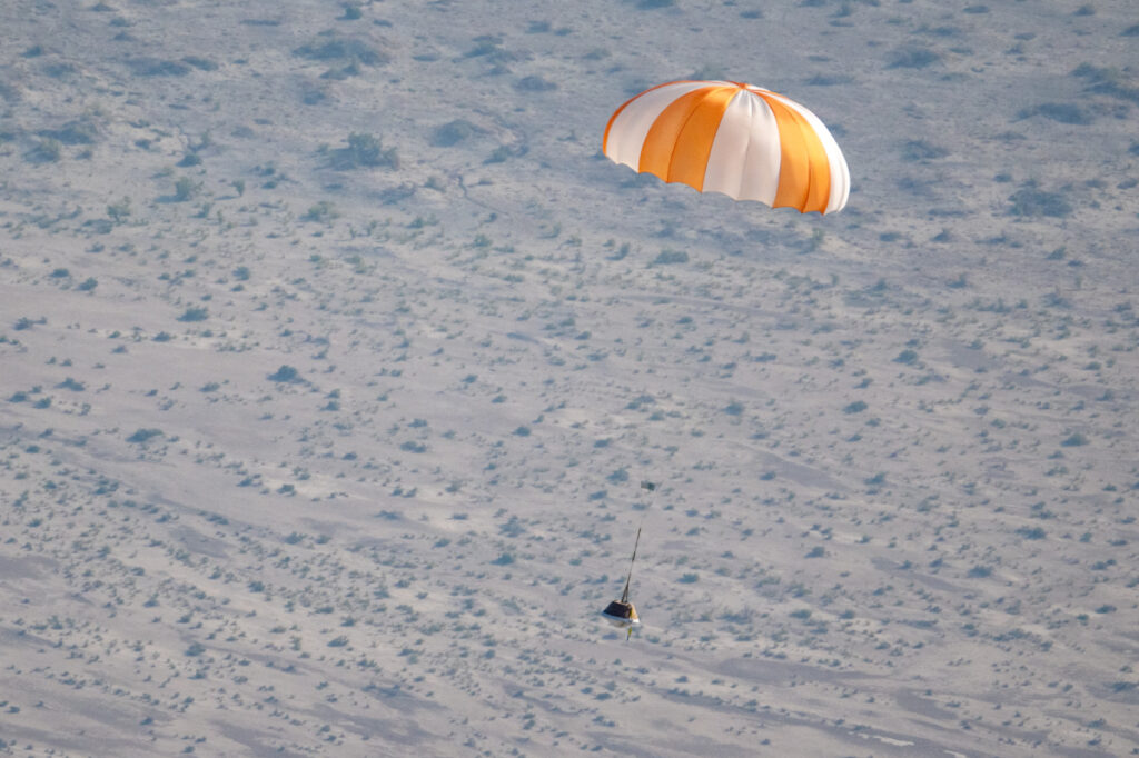 A tan and white diamond-shaped capsule descends toward the desert floor suspended from a white and orange parachute.