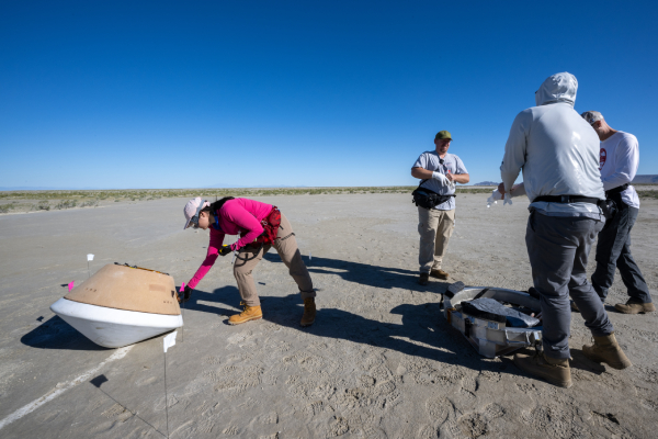 A person in a pink shirt places flags around a tan and white model of the sample return capsule as it sits on the desert floor. Three other scientists in white don protective gear next to the capsule.