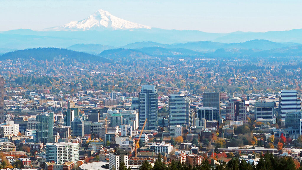 In the background, snow-capped Mount Hood rises above a mountain range. In the foreground, the skyscrapers of Portland, Ore., dominate the landscape.