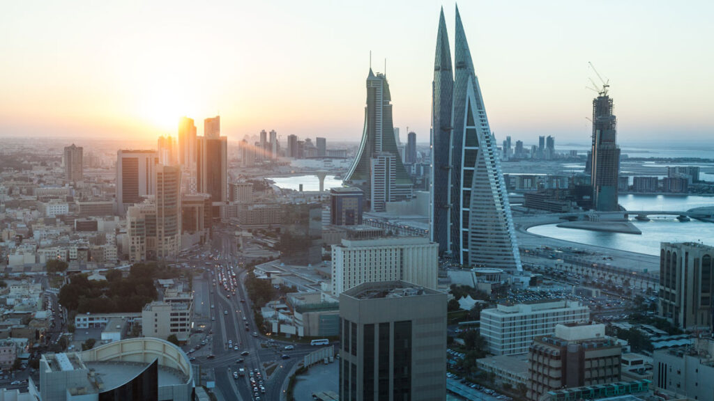 Tall buildings, including several shaped like triangles, are densely packed together, and the Sun appears on the horizon in the background.