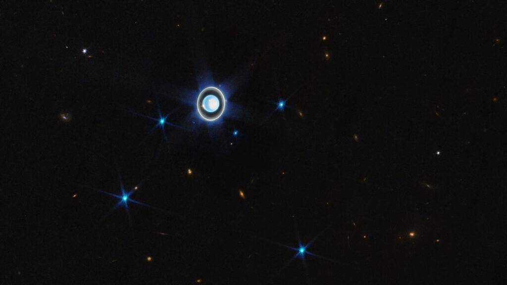 Uranus is a shiny blue-white orb, slightly off center in this image. Its rings are seen nearly face on, and six bright blue dots show the six brightest moons of the planet. There are several reddish galaxies in the background.