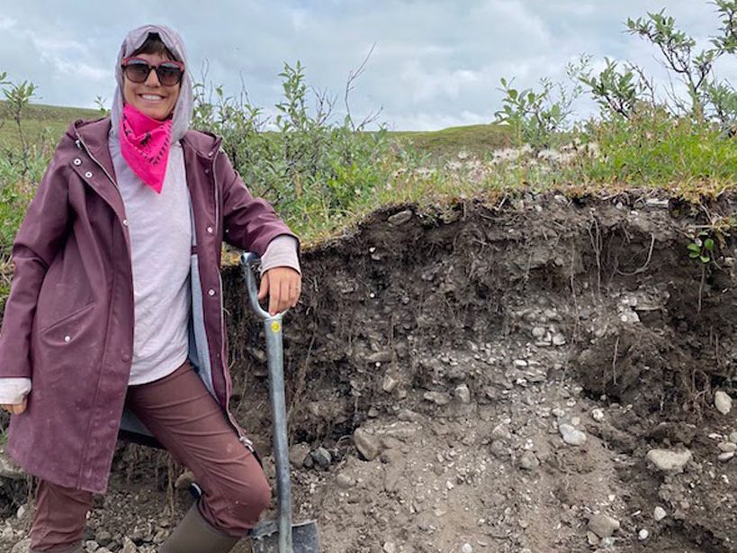 A person wearing sunglasses is looking at the camera and leaning on a shovel next to an outcrop of soil. A green landscape is in the background.