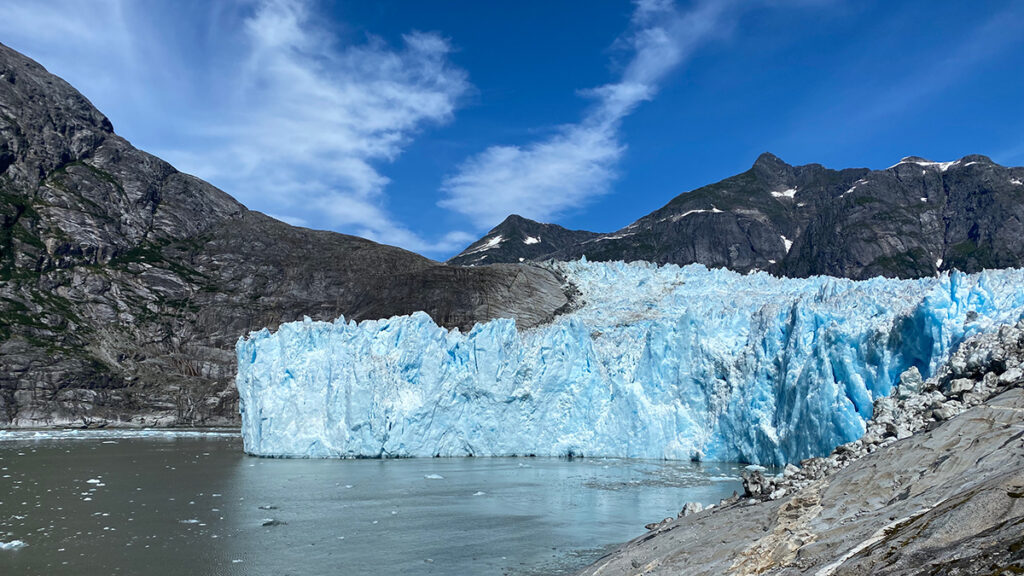 Jagged-surfaced blue-white glacier, surrounded in the foreground by seawater and in the background by dark colored, snowcapped mountains
