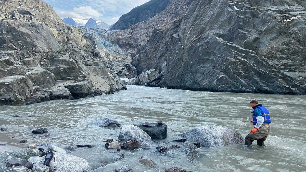 A man wearing a blue life vest stands knee deep in water to collect a water sample downstream of the large glacier in the background.