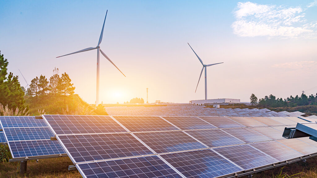 Solar panels in the foreground in front of two wind turbines, with the Sun low on the horizon in the distance