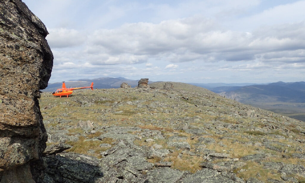 An orange helicopter sits on a rocky hilltop amid a vast undeveloped landscape.