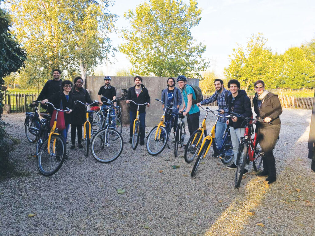 Ten people standing next to their bicycles in a semicircle. Trees are in the background, and there’s gravel on the ground.