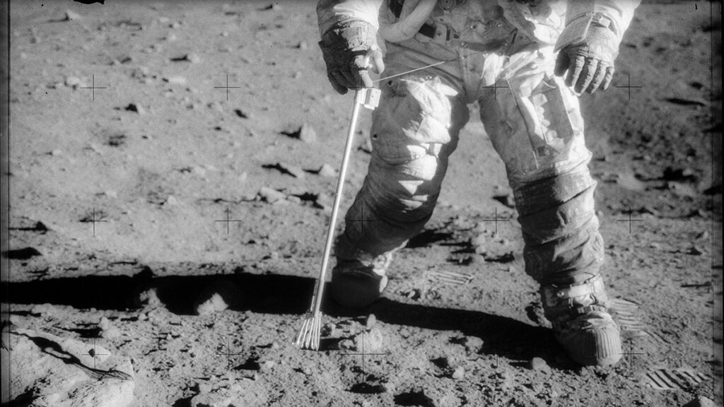 A view of the lower body of an astronaut carrying tongs and walking on the surface of the Moon