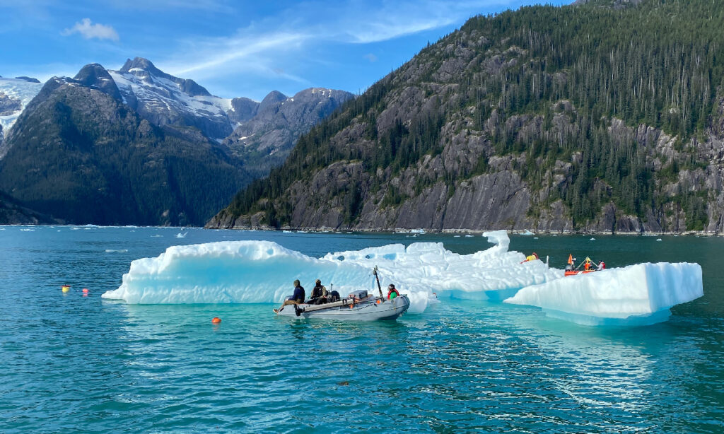 Grupo de científicos en una pequeña embarcación, investigando una balsa de hielo glaciar que flota en un mar turquesa. Al fondo, escarpadas montañas nevadas.