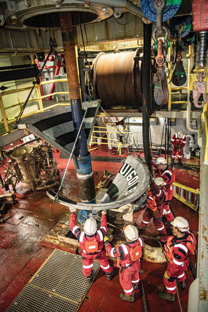 Several people, dressed in red uniforms and white hardhats, surrounded by pipes and machines.